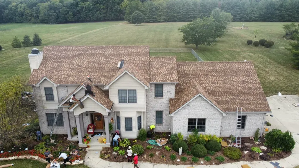 An aerial view of workers repairing the shingled roof of a two-story brick house surrounded by a rural landscape.