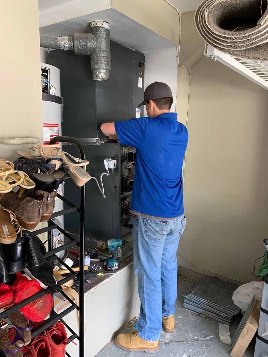Technician in blue shirt inspecting HVAC unit in a utility closet, with tools and shelving nearby