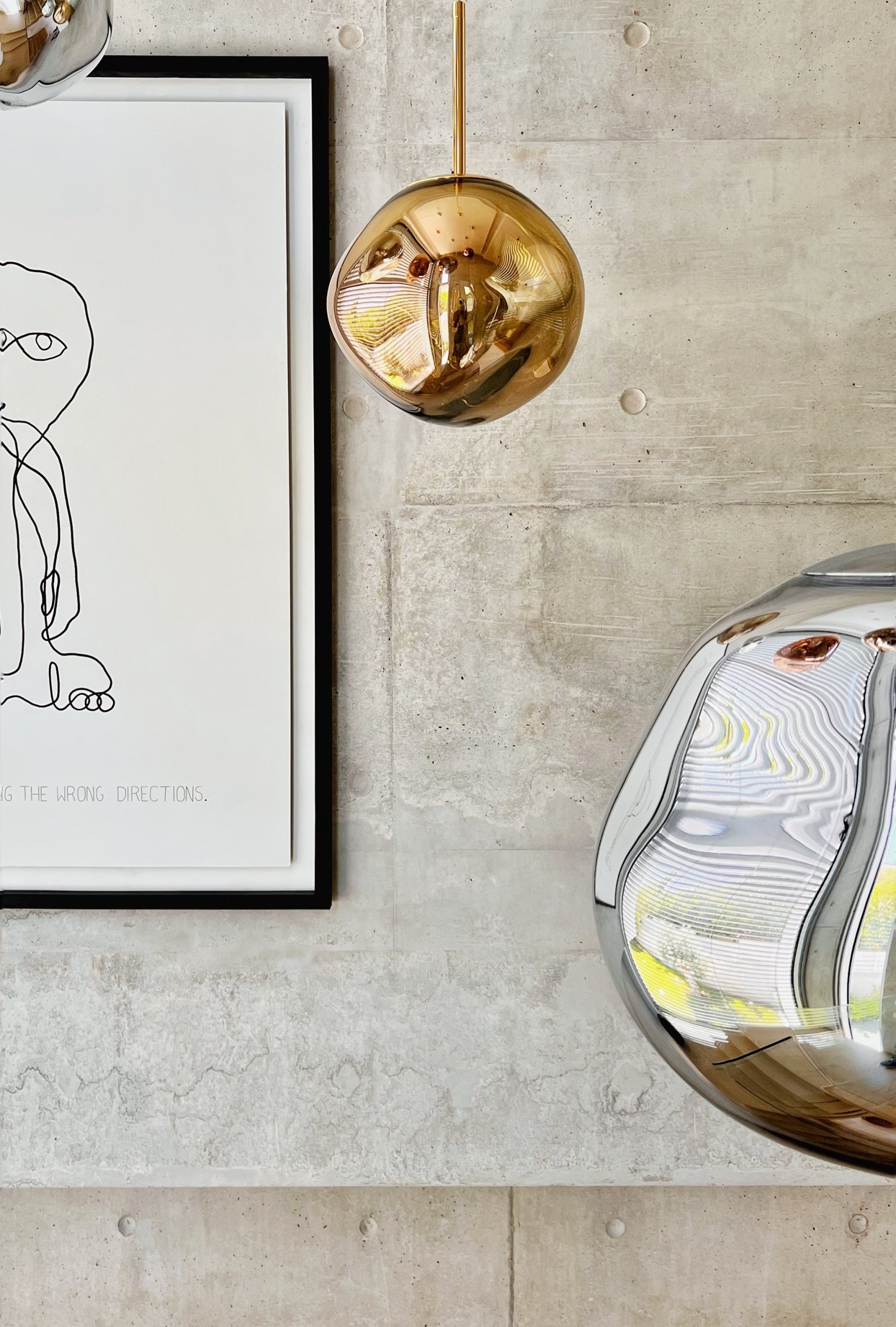 Metallic gold and silver pendant lights hang against a concrete wall, near a framed black-and-white drawing.