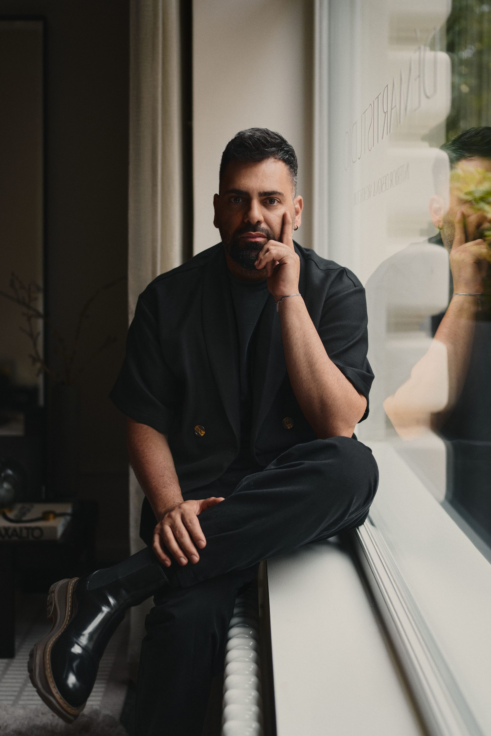 Elegant man in black sitting on a window bench inside a luxury interior designed by DÉMARTISTUDIO, surrounded by warm light, architectural details, and contemporary furniture.