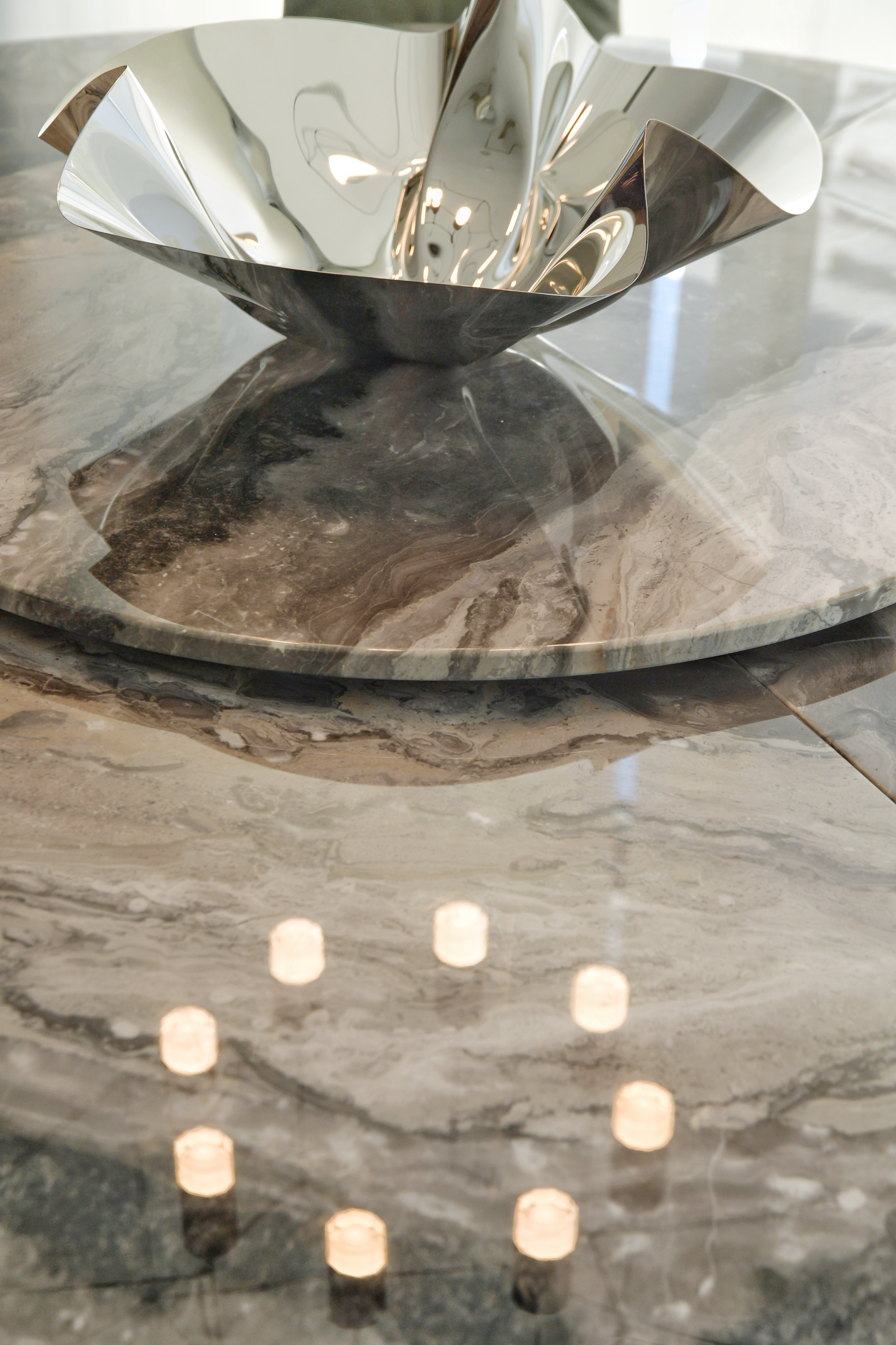 A shiny silver bowl on a marble table, with a reflection of lights beneath it.