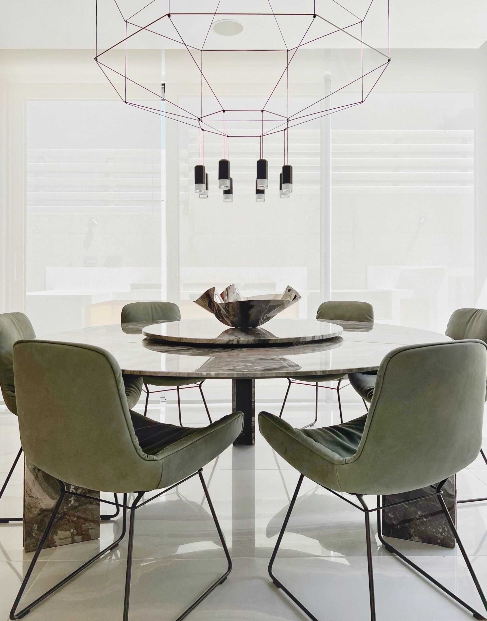 Modern dining room with round marble table, green chairs, and geometric chandelier.