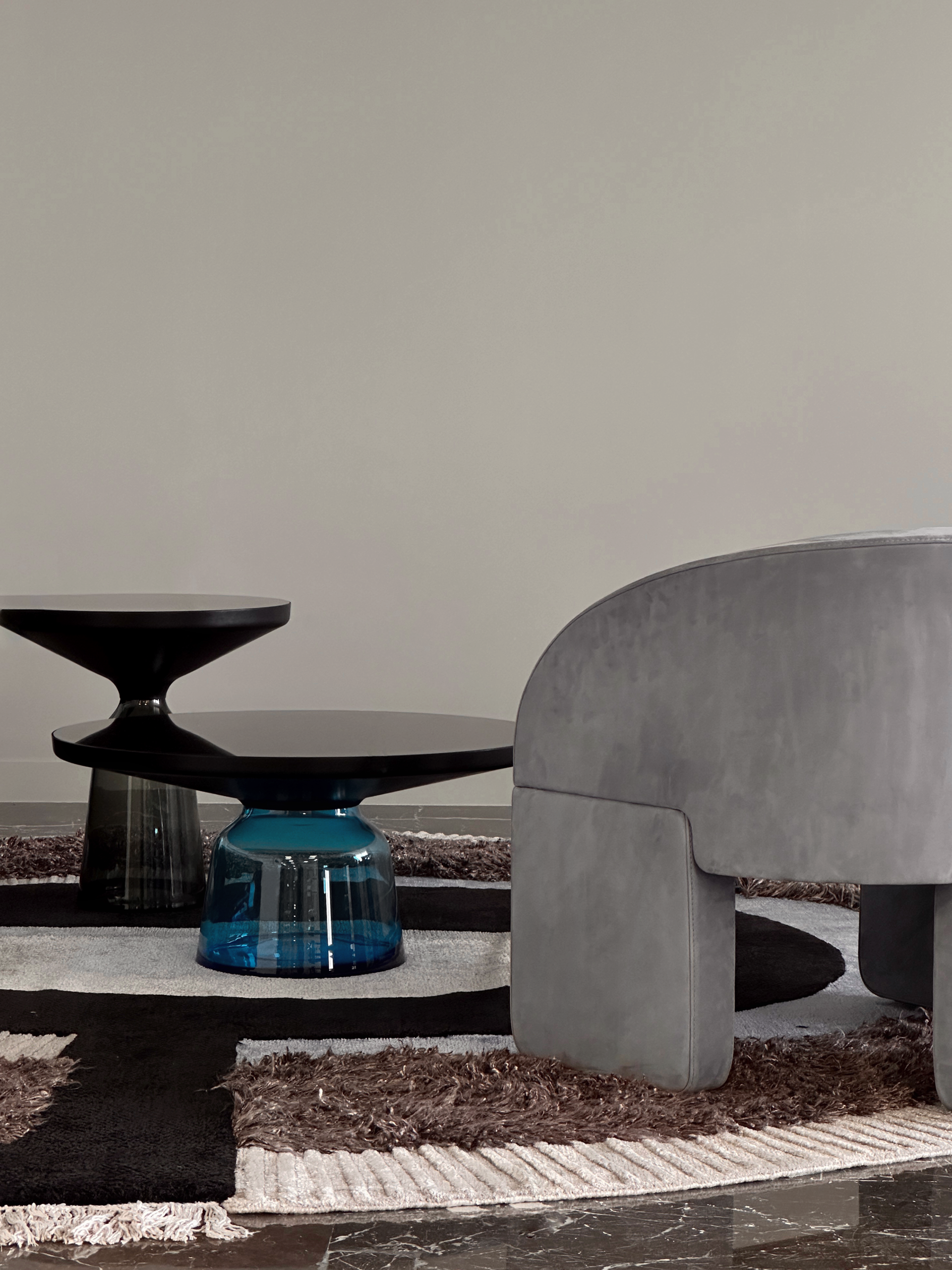Two modern side tables, one black, one blue glass, on a rug next to a gray curved sofa.