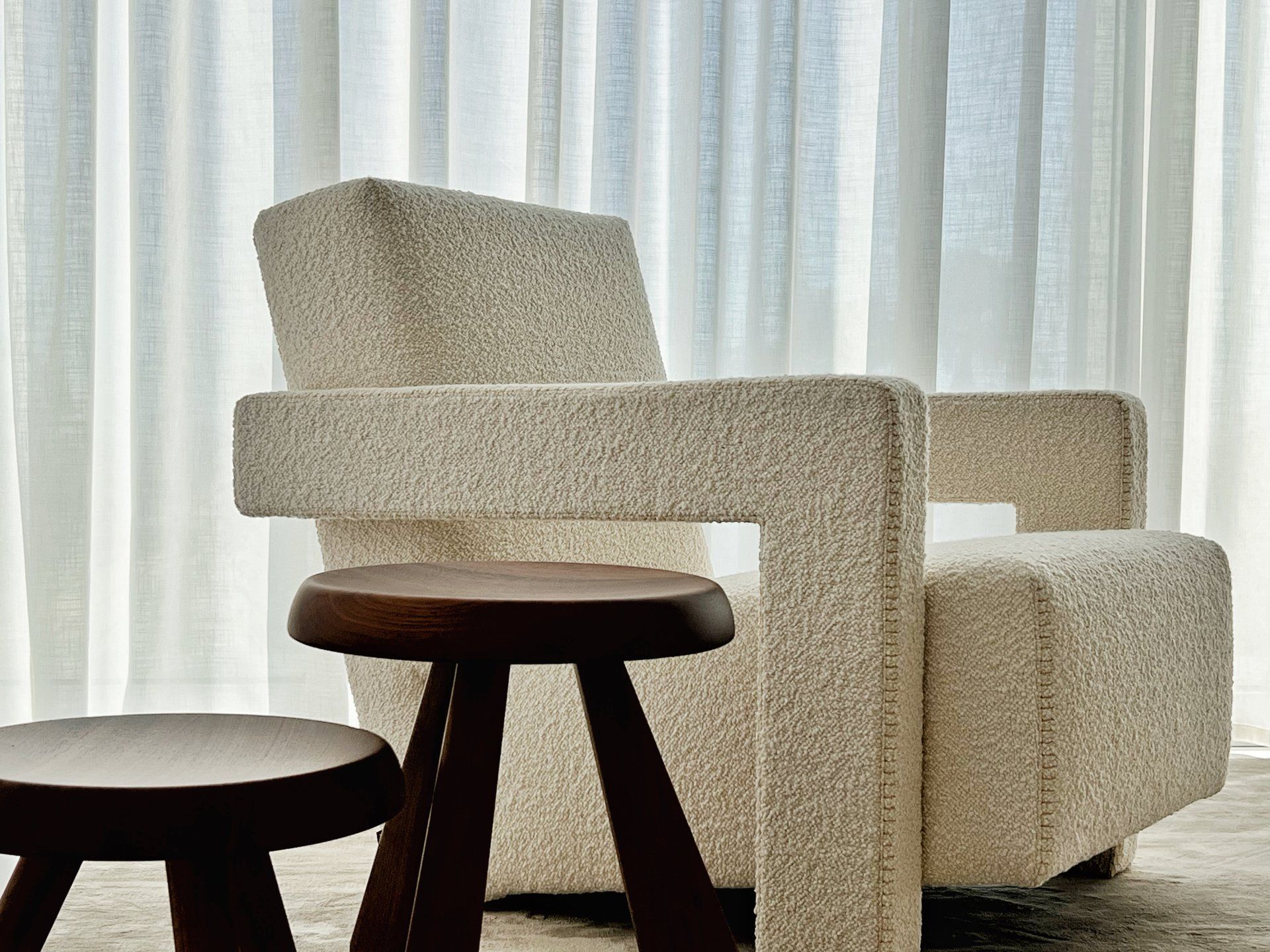 Cream-colored boucle armchair and dark wooden stool in front of a sheer white curtain.