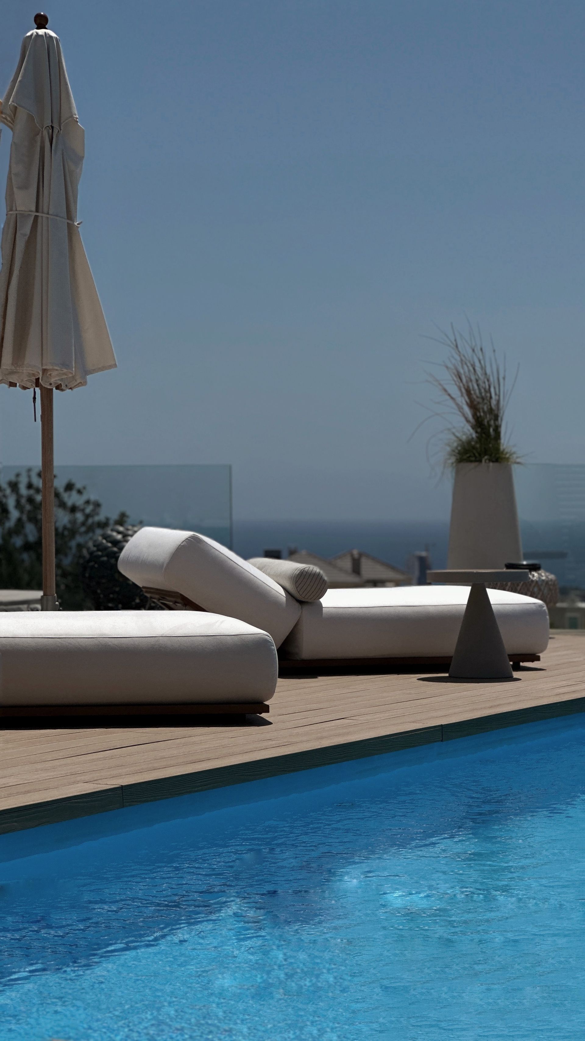Poolside scene with white lounge chairs and umbrella, blue water, clear sky.