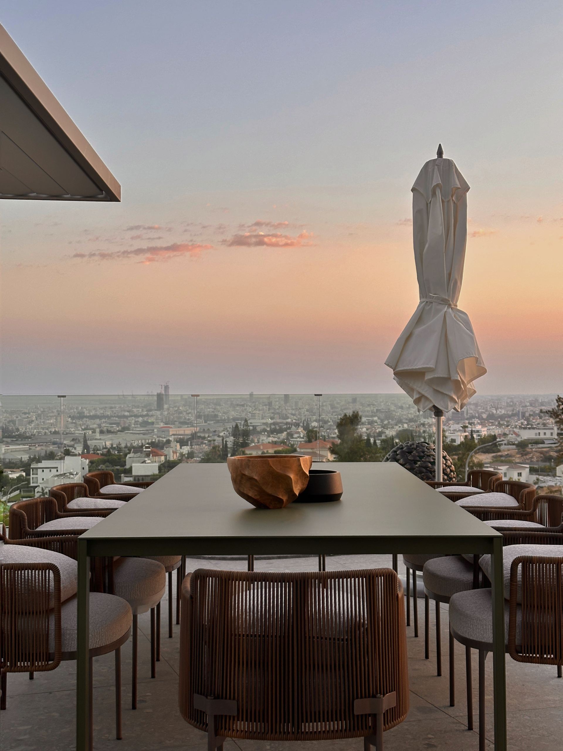 Outdoor dining table and chairs overlooking a city at sunset, with a closed umbrella.
