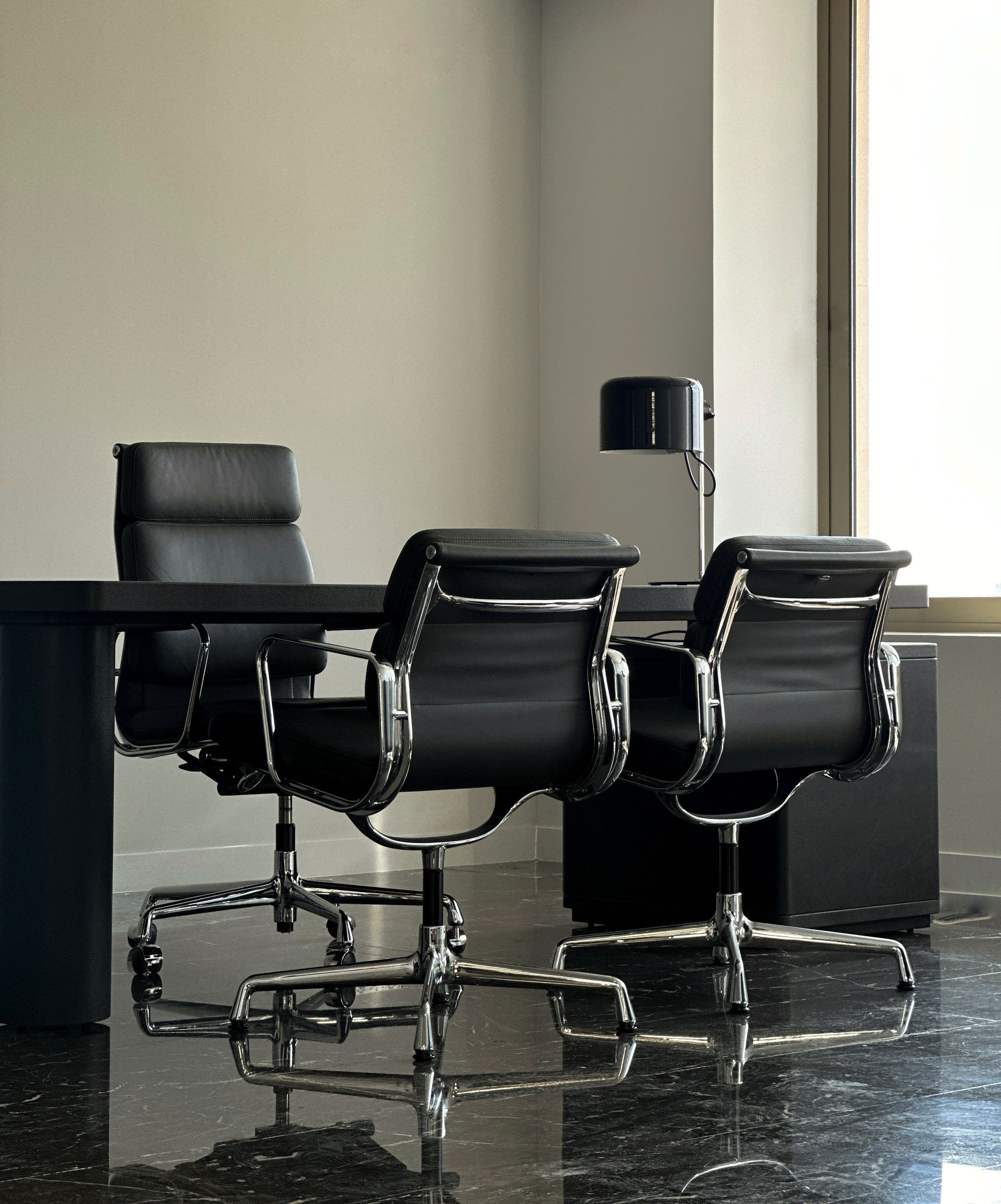 Black leather office desk with chairs, lamp, and window. Reflective flooring and light walls.