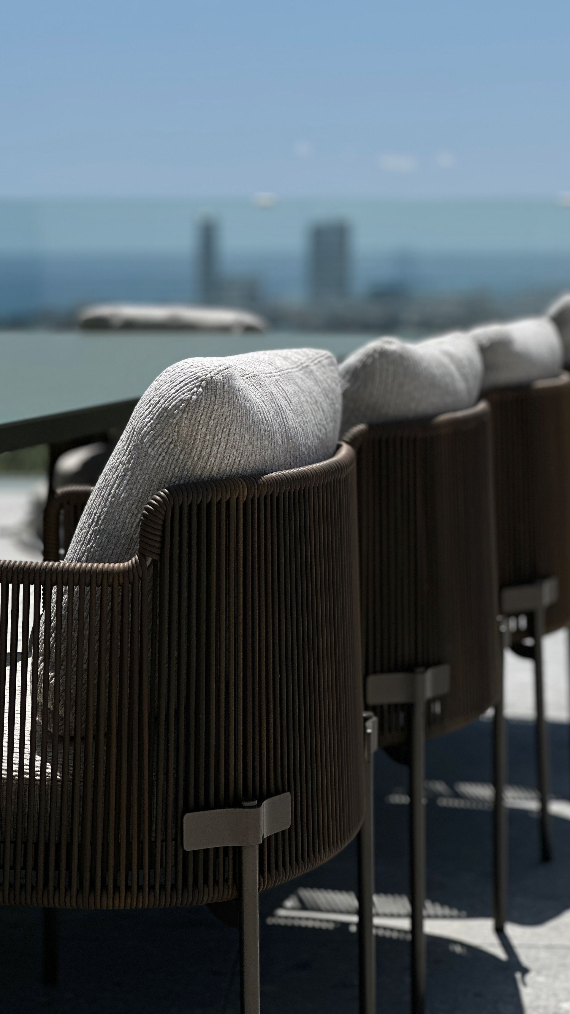 Rope-backed chairs with white cushions on a terrace overlooking water and city buildings on a sunny day.