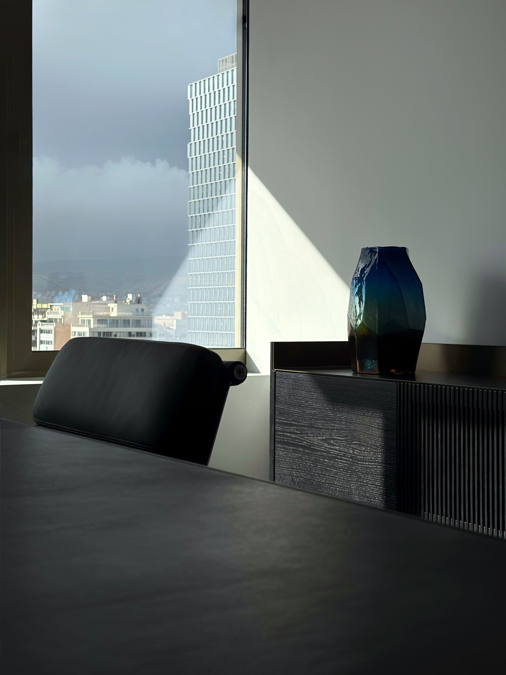 Modern room with blue vase, black chair, window with a skyscraper, and a dark table.