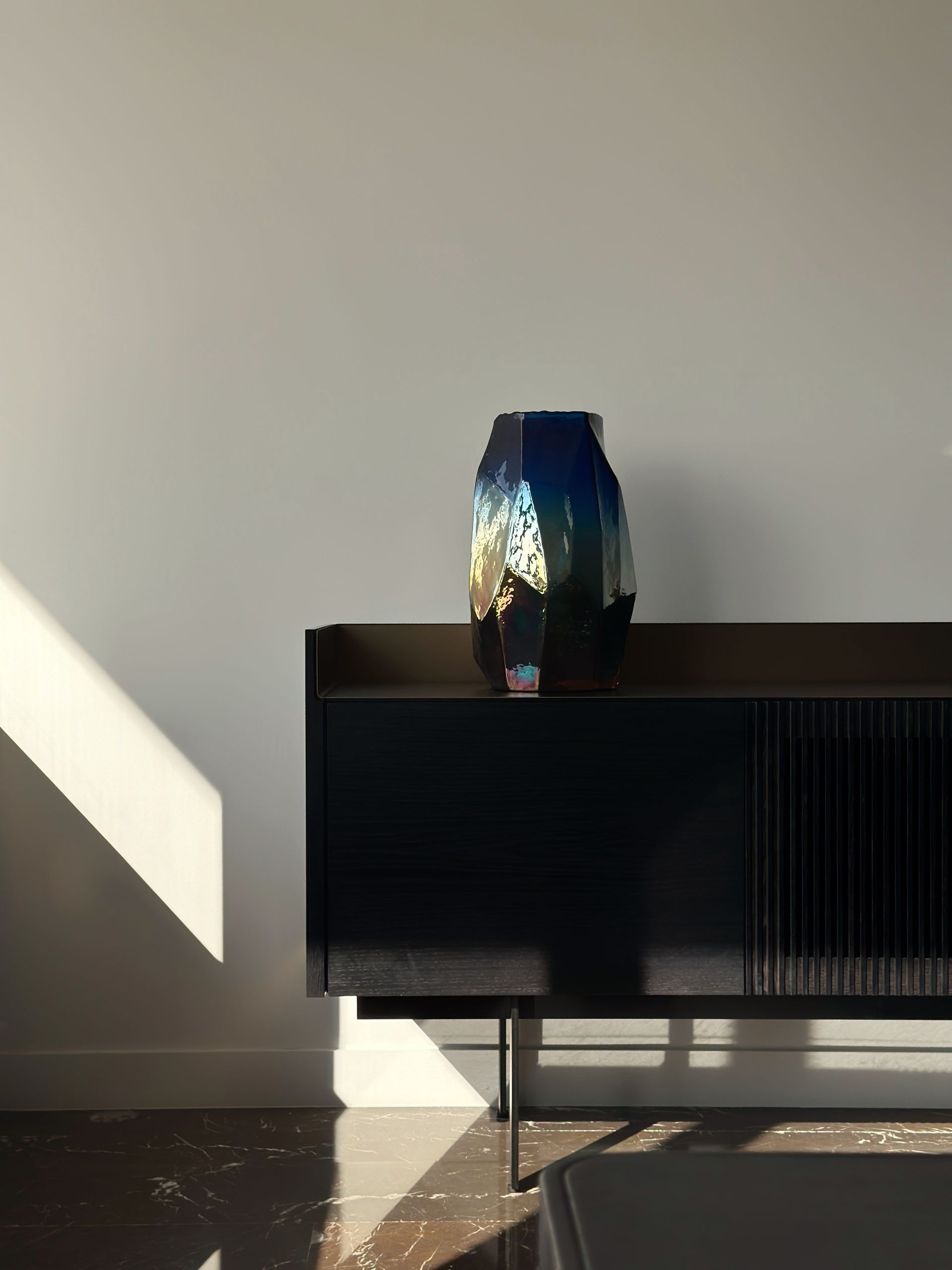 Blue and gold textured vase on a dark cabinet against a white wall with sunlight.