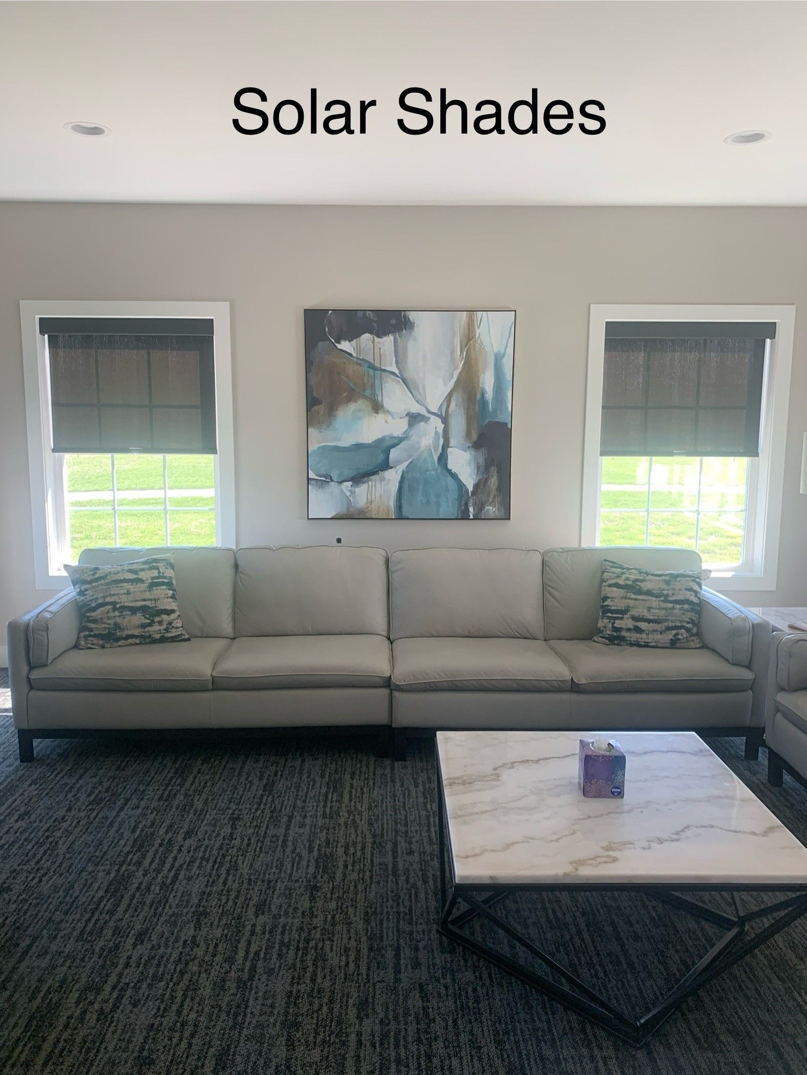 A living room with a couch , coffee table , and two windows with solar shades.