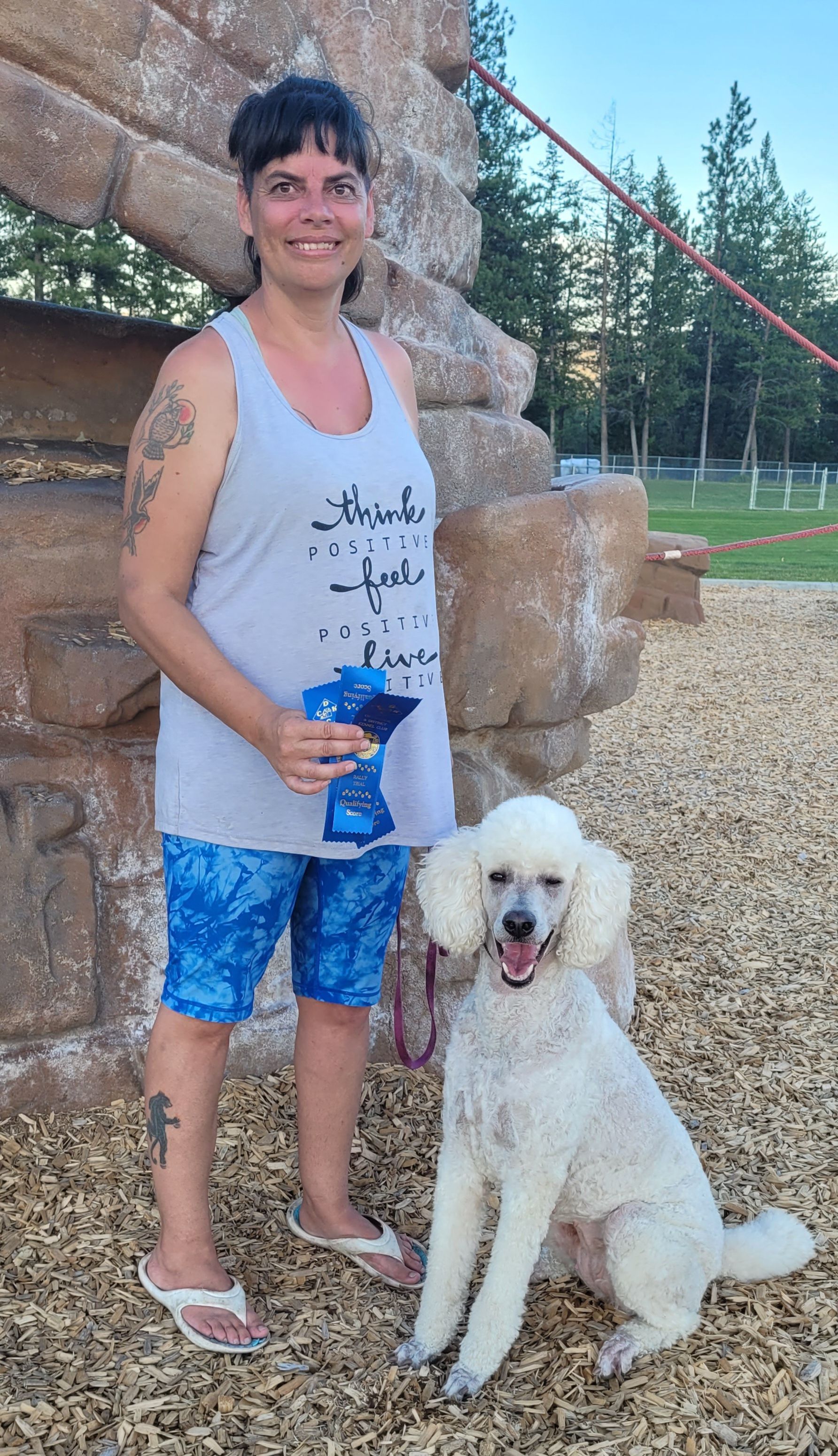 A woman with tattoos on her arm and leg stands on wood chips next to a white standard poodle in an outdoor setting.