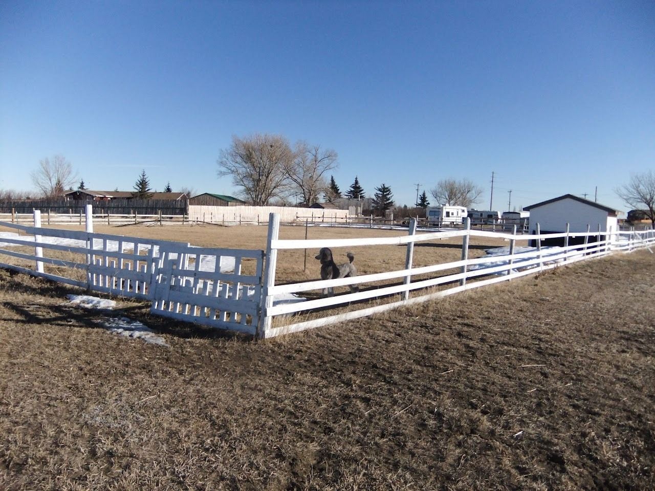 White fence encloses a grassy area with a building. A few trees and a clear blue sky are visible.