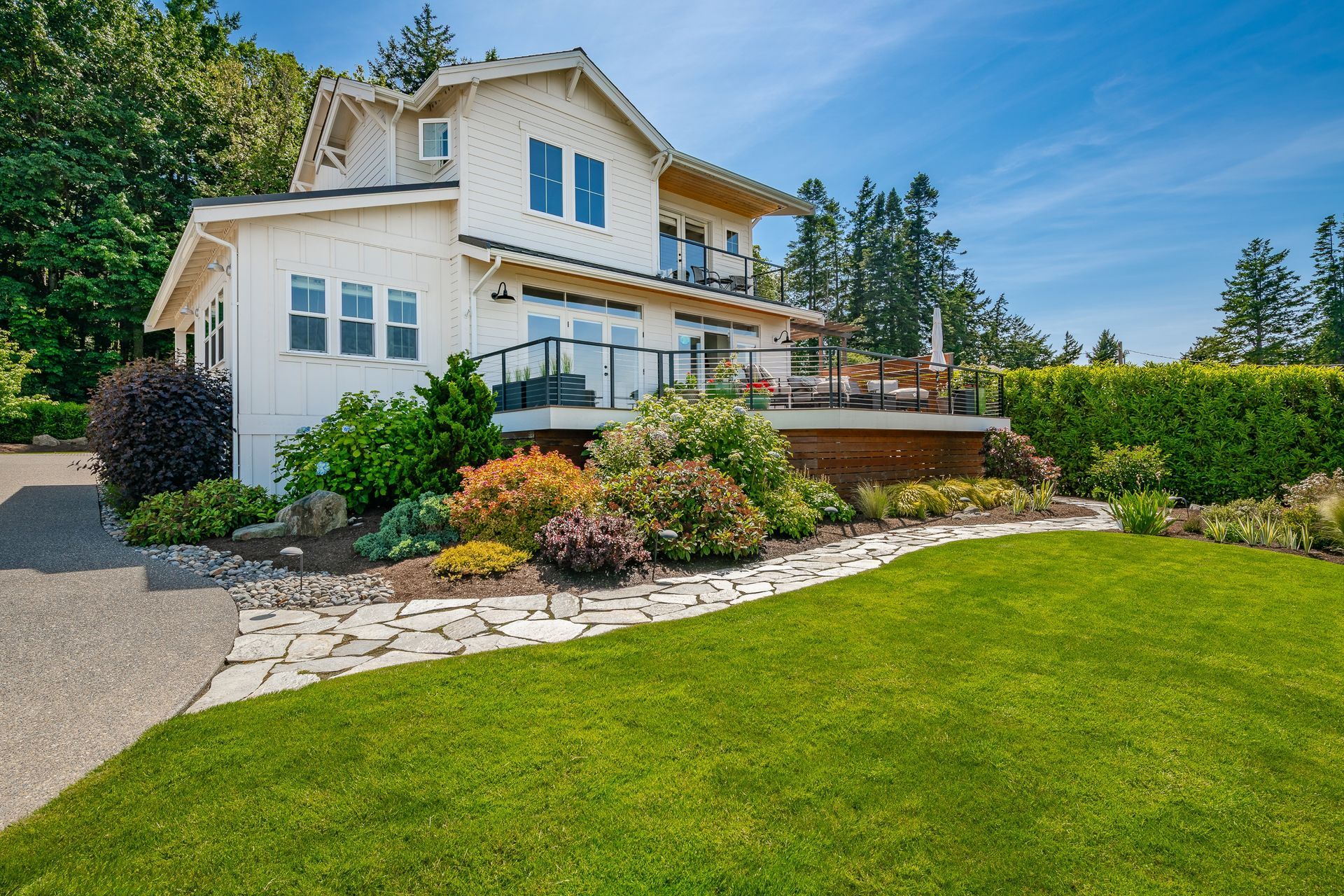 Two-story beige house with a manicured lawn, flower beds, and a stone walkway under a blue sky