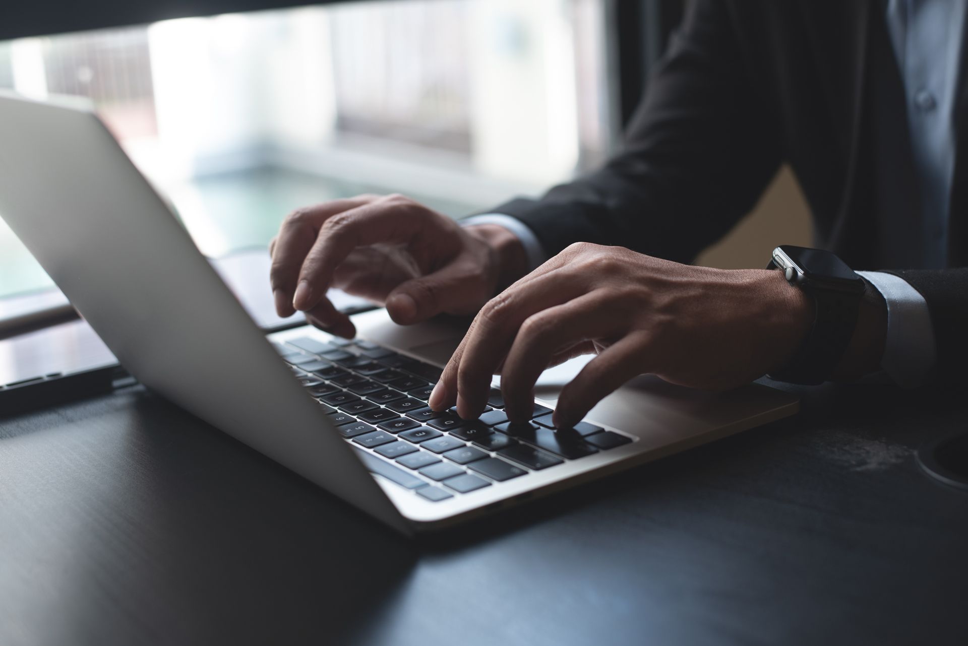 Person in a suit typing on a laptop at a desk near a window.