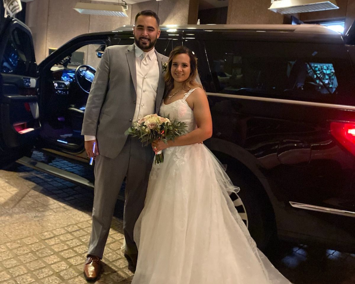 A bride and groom are standing in front of a black suv.