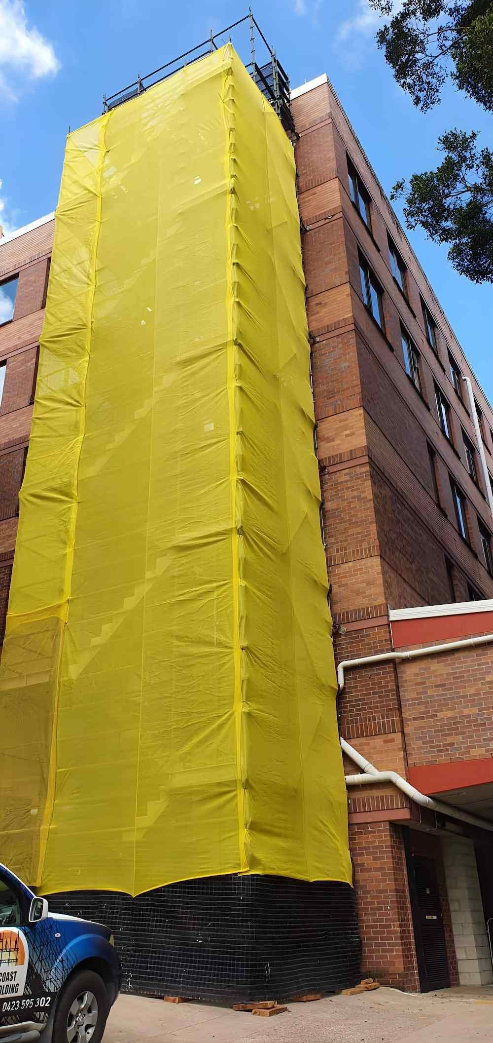 A Large Brick Building is Covered in Yellow Tarps — All Coast Scaffolding In Yandina, QLD