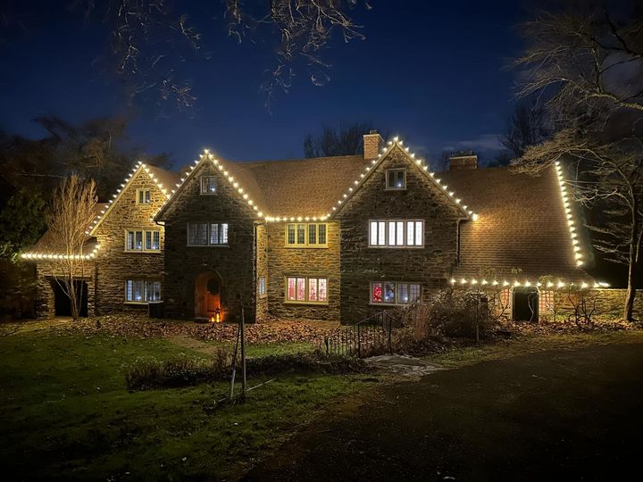 Large stone house at night with string lights along the roofline; lit windows and dark trees in the background.