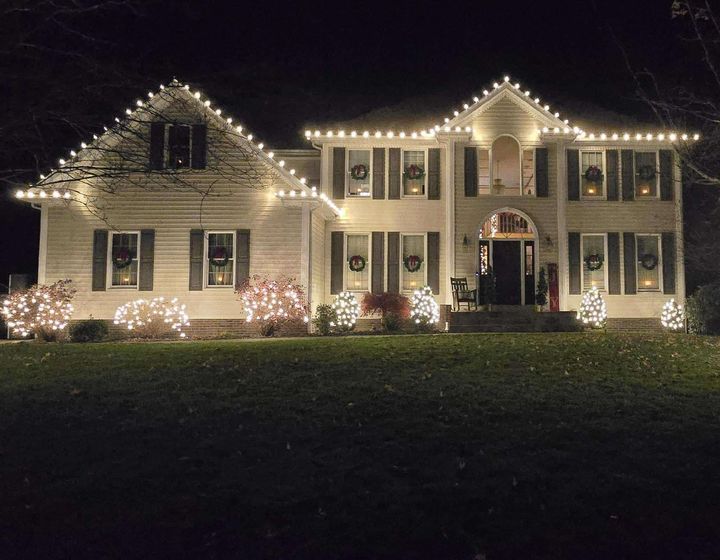White house decorated with Christmas lights, wreaths, and bushes illuminated at night.