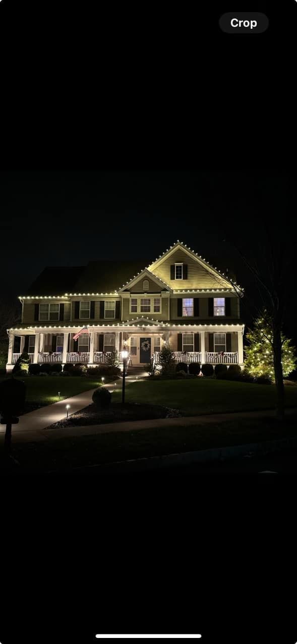 A large house at night with Christmas lights on the roof and porch, surrounded by illuminated landscaping.