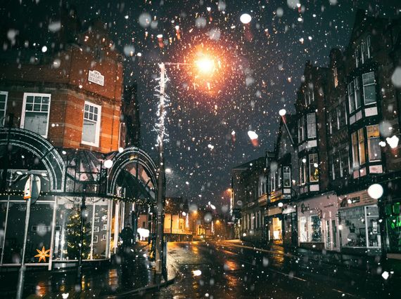 Snowy street scene at night, with brick buildings and streetlights. Snow falls over the wet pavement.