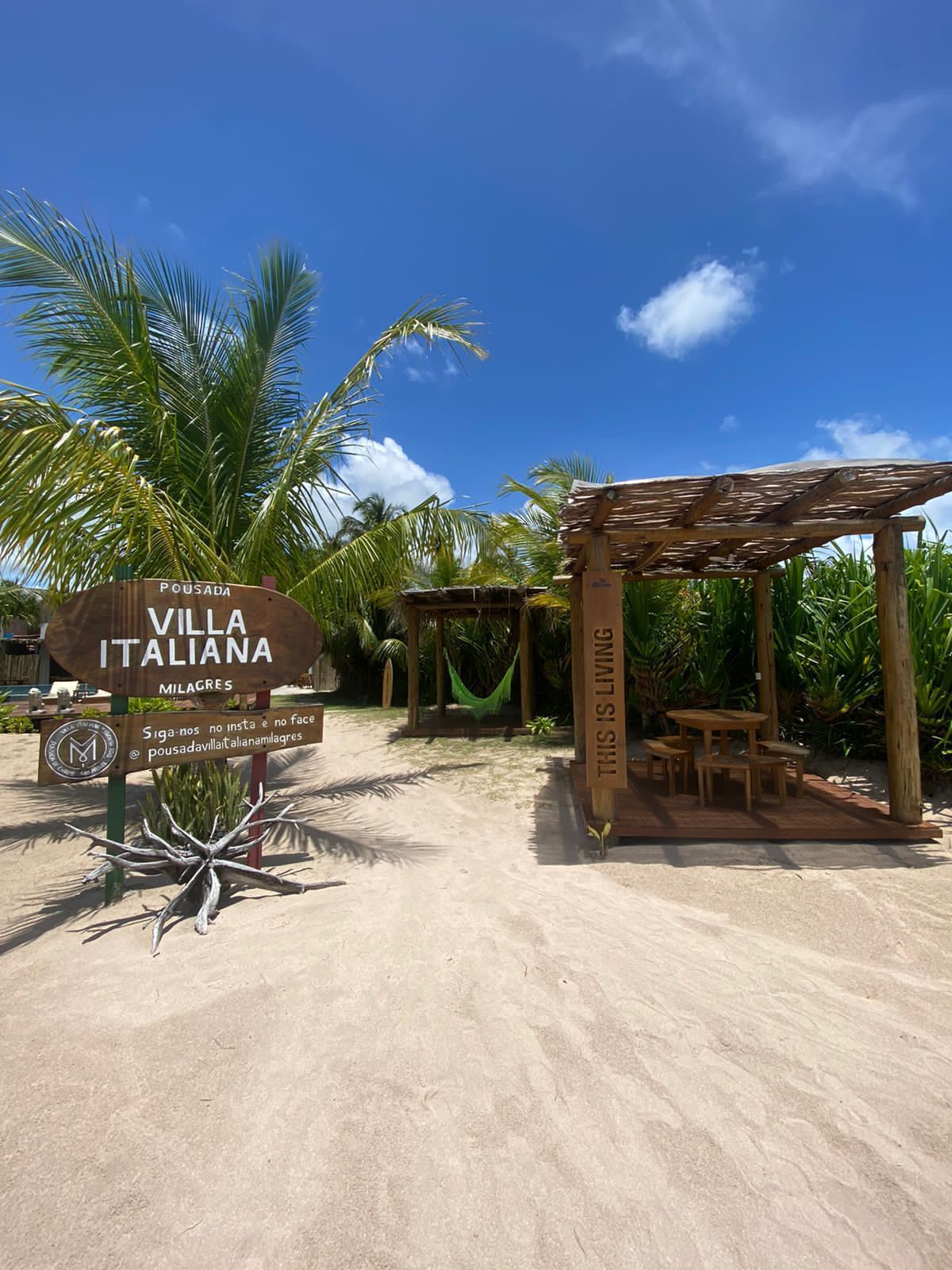 Placa da Villa Italiana e estruturas de madeira na praia de areia sob o céu azul.Pousada Villa Italiana