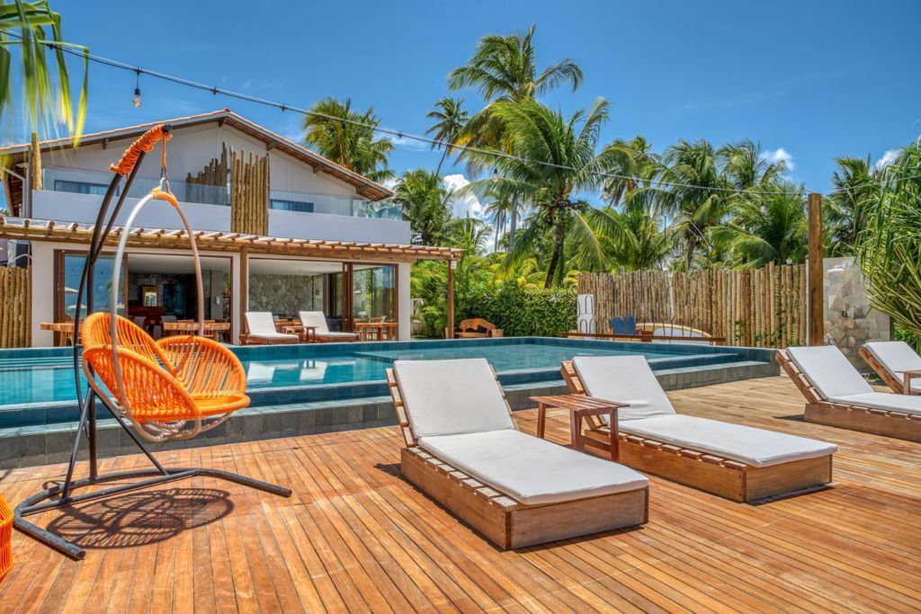 Um resort com piscina e espreguiçadeiras; balanço laranja; céu azul e palmeiras. Pousada Villa Italiana