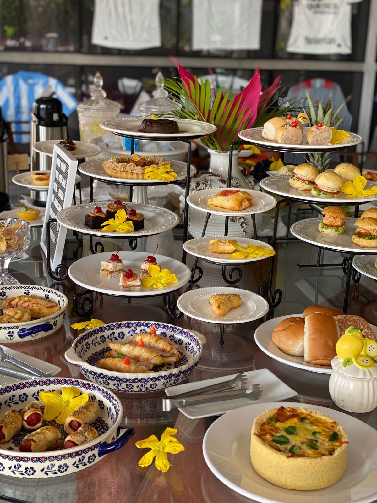 Mesa de bufê com diversos doces, bolos e salgados. Flores coloridas e pratos em camadas.