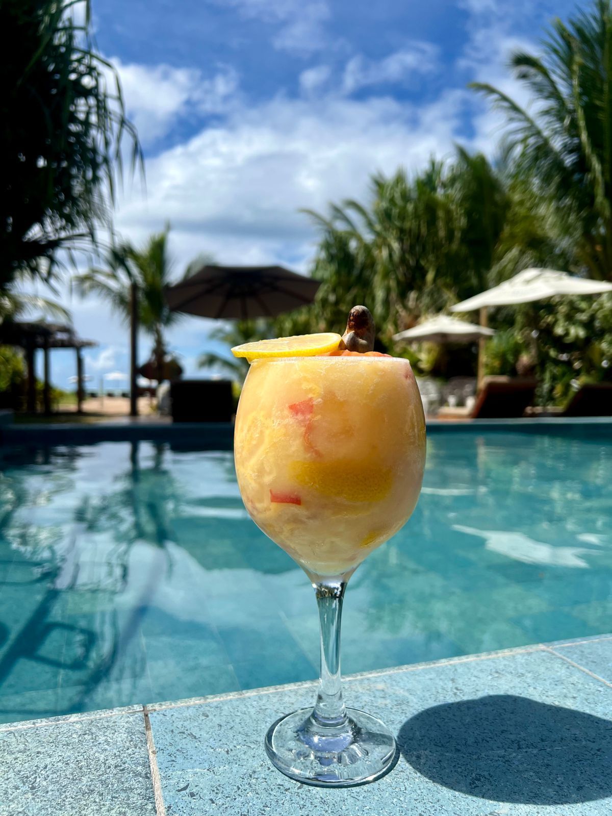 Um coquetel cor de pêssego em um copo à beira da piscina com vista para a praia sob um céu azul.Pousada Villa Italiana