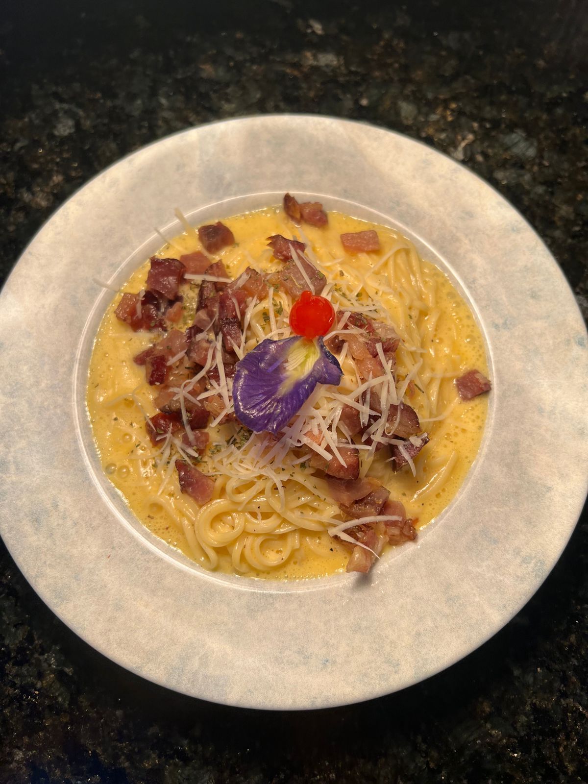 Prato de massa com bacon, queijo e uma guarnição de flores roxas em uma tigela branca.Pousada Villa Italiana