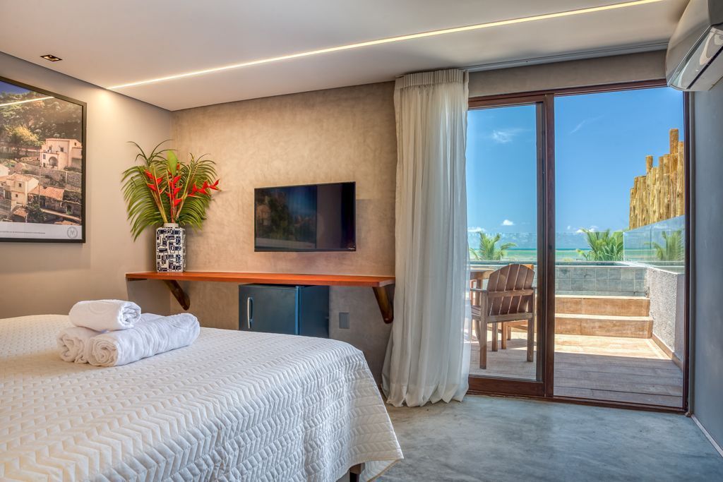 Quarto com vista para o mar, cama branca, TV e porta de vidro deslizante para um pátio.Pousada Villa Italiana