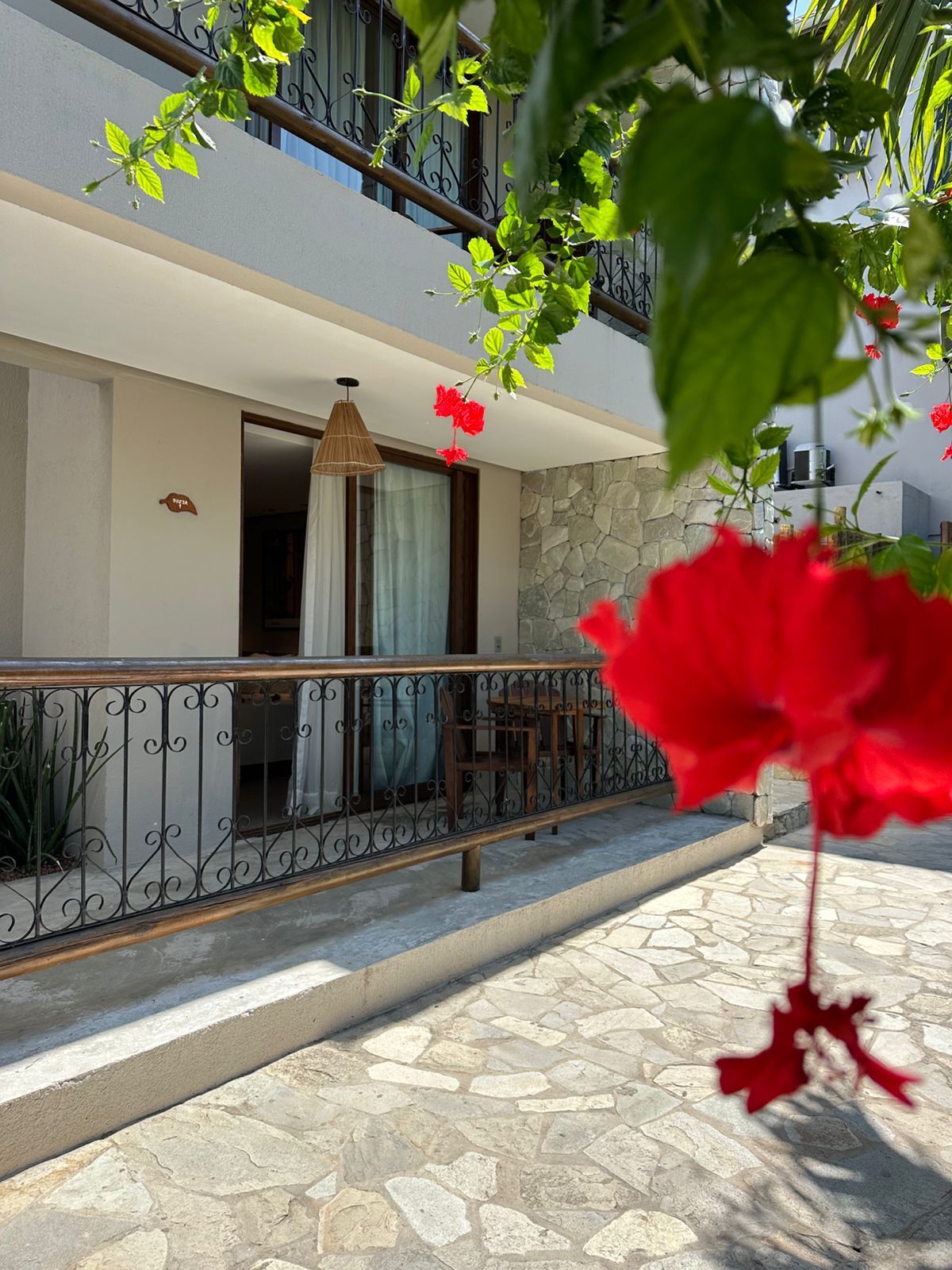 Varanda com corrimão ornamentado, flor de hibisco vermelho em primeiro plano, paredes bege, porta aberta.Pousada Villa Italiana