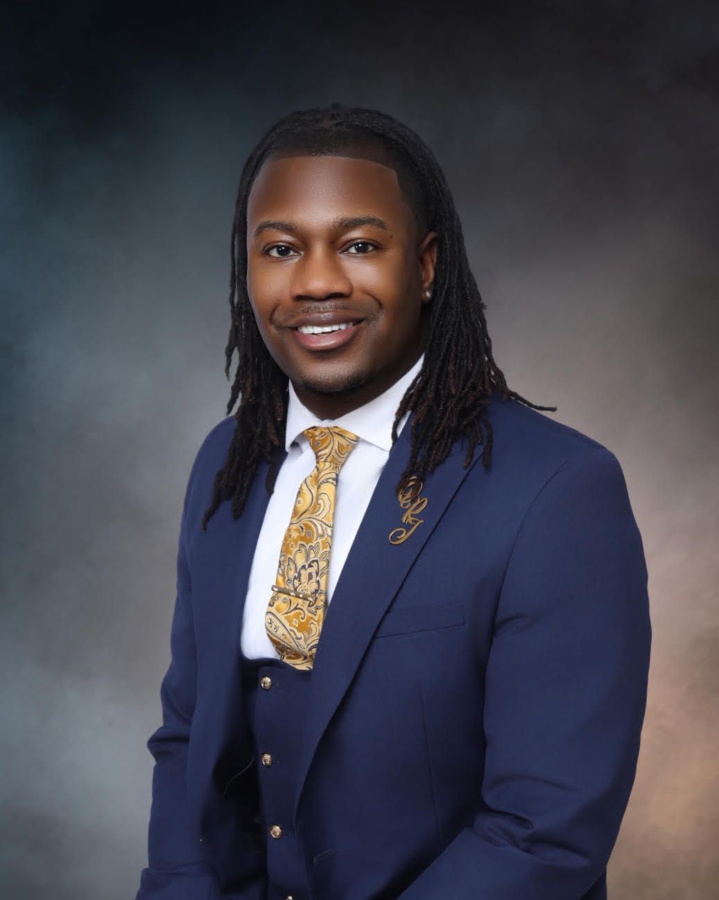 Studio portrait of a smiling person in a navy suit with a gold tie against a gray backdrop