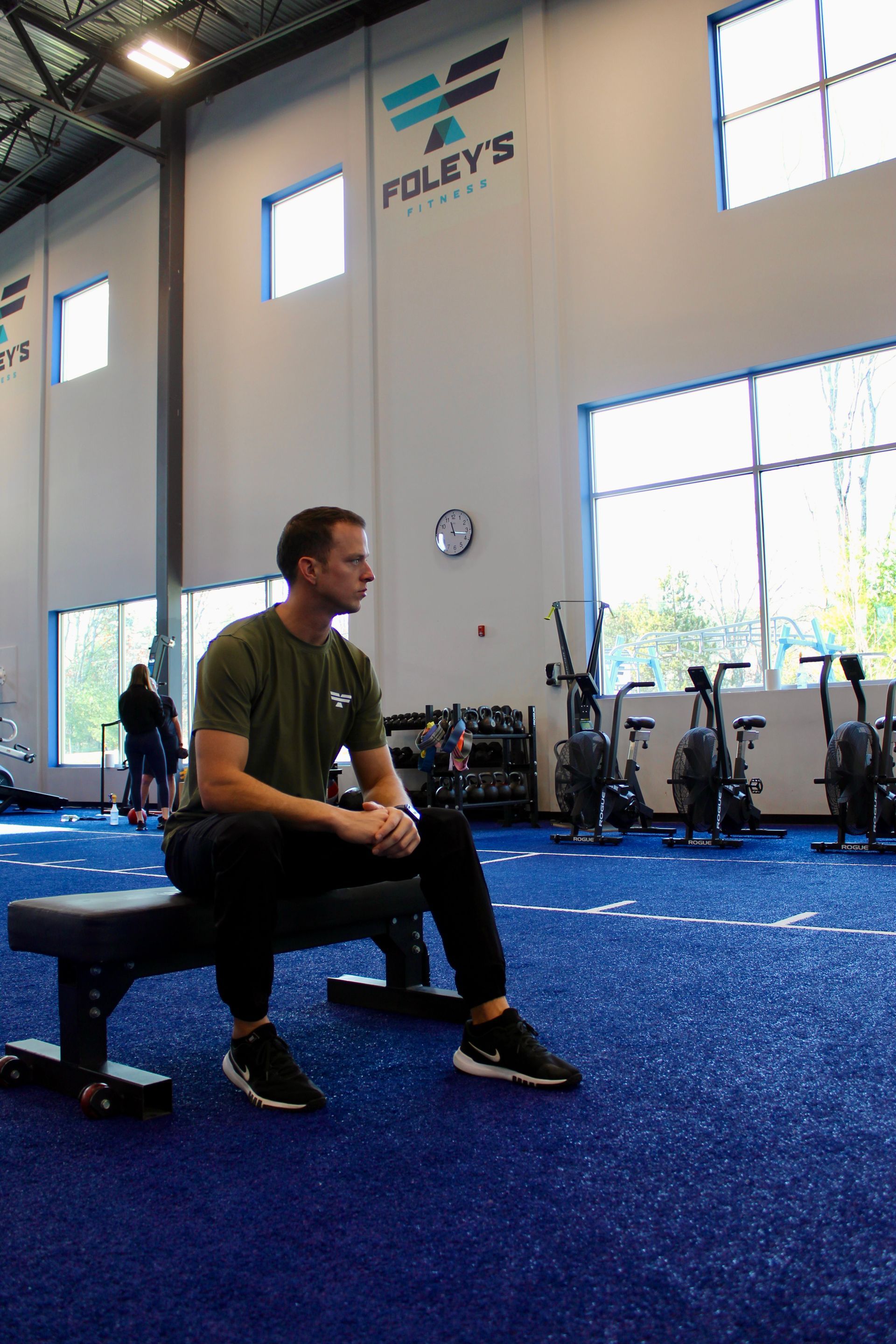Man in olive shirt sits on a bench in a gym, looking to the side. Blue floor, white walls.