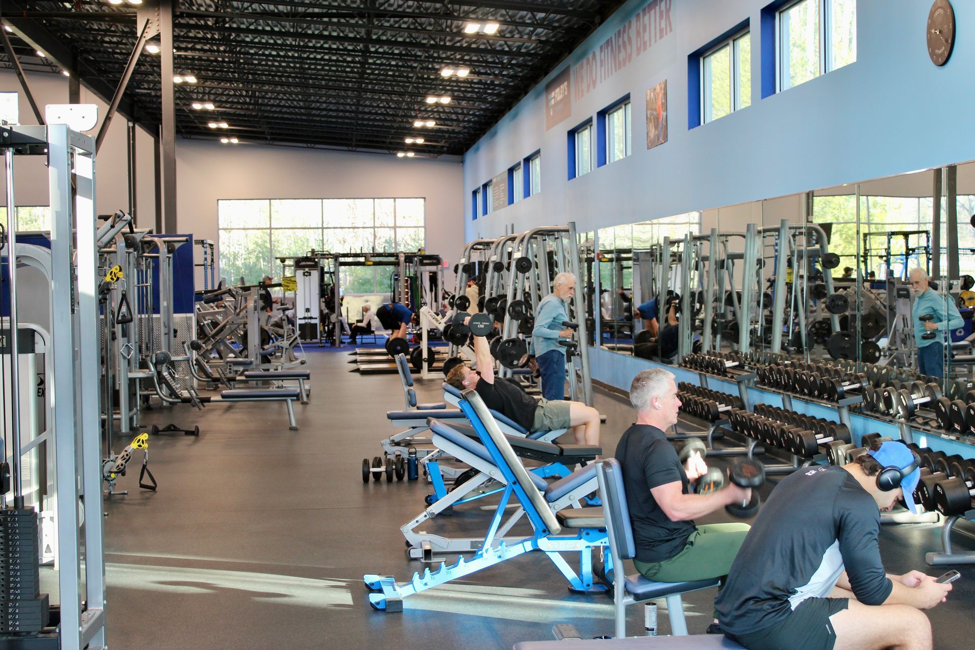 Inside a gym with people using weight machines and free weights. Mirrors, windows, and blue and grey equipment.