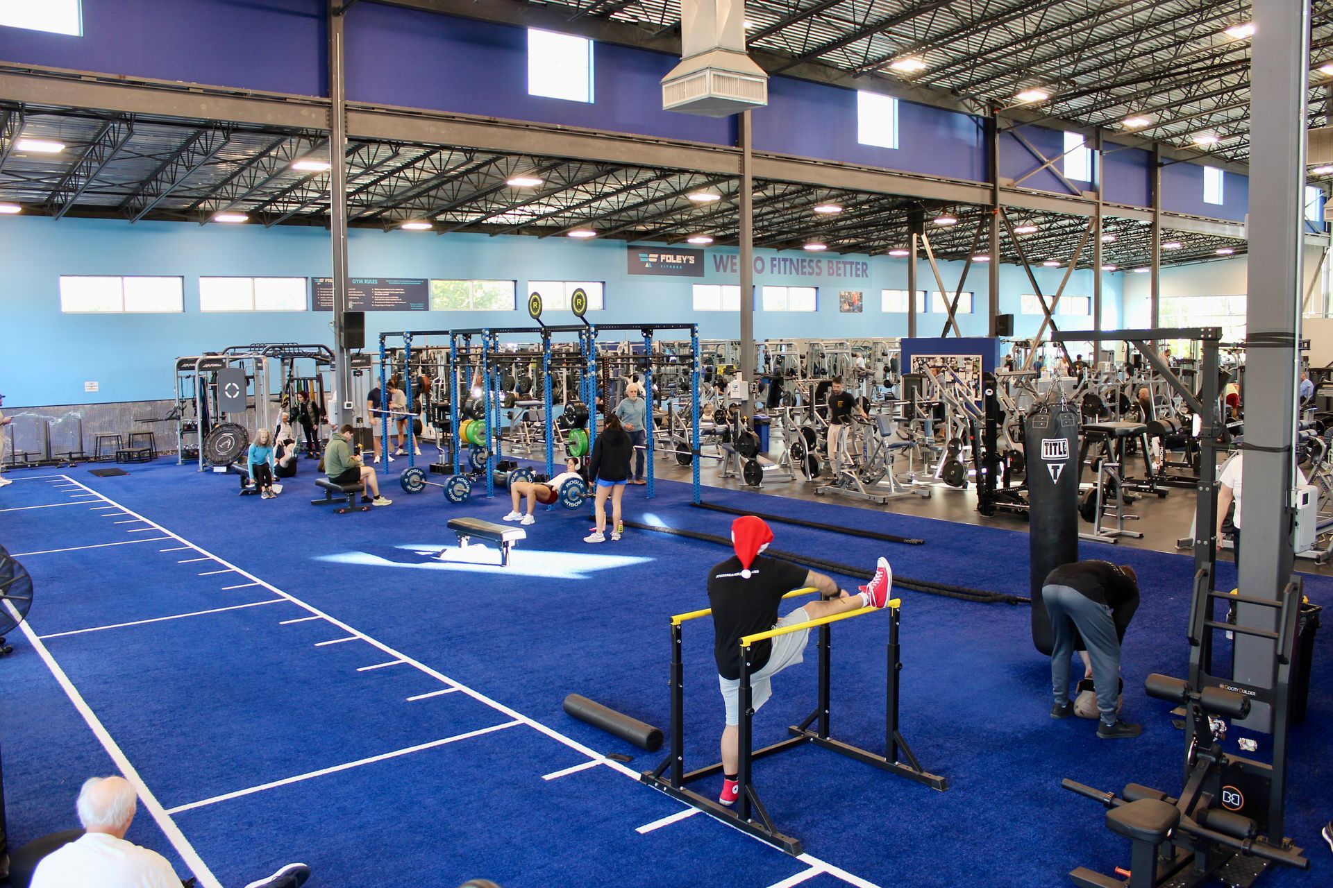 Blue-floored gym interior with fitness equipment and people exercising. Some stretching, others using machines.