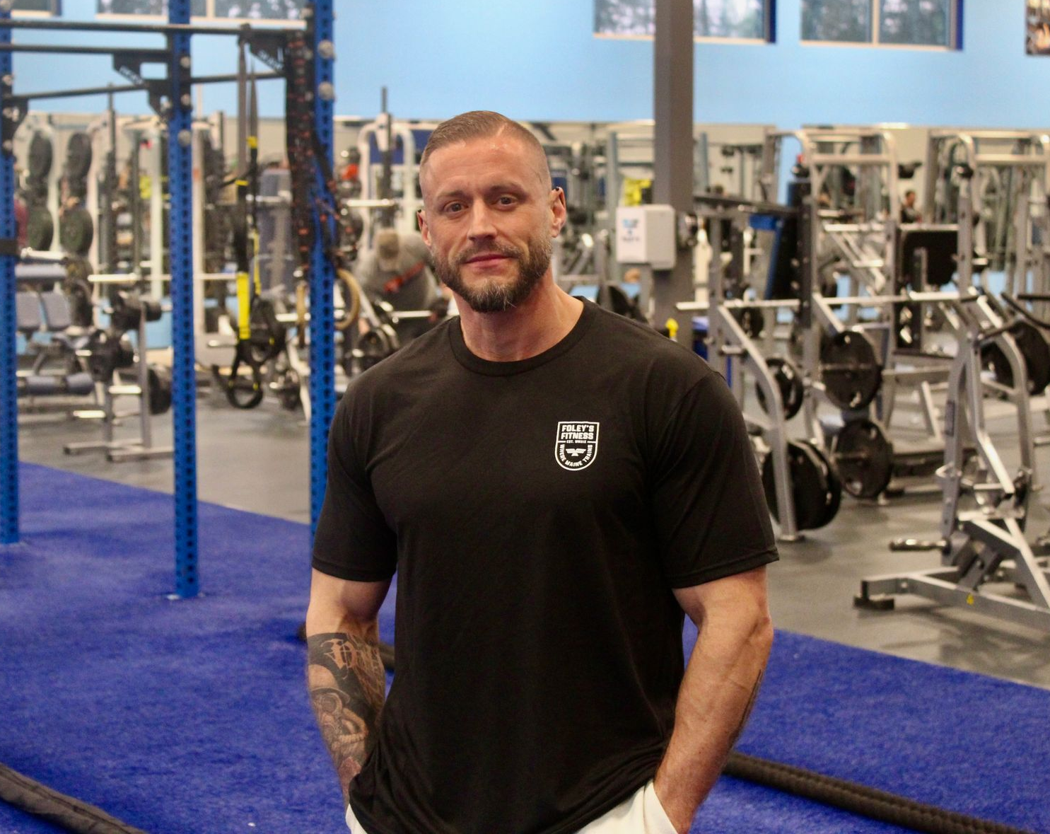 Man in black shirt stands in a gym, arms at his sides, blue and silver equipment in background.
