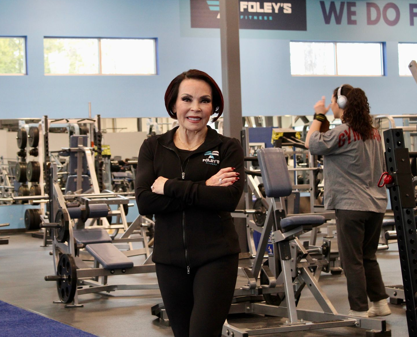 Woman in a gym, arms crossed, smiling.  Gym equipment in background. Another person wearing headphones. Foley's Fitness sign.