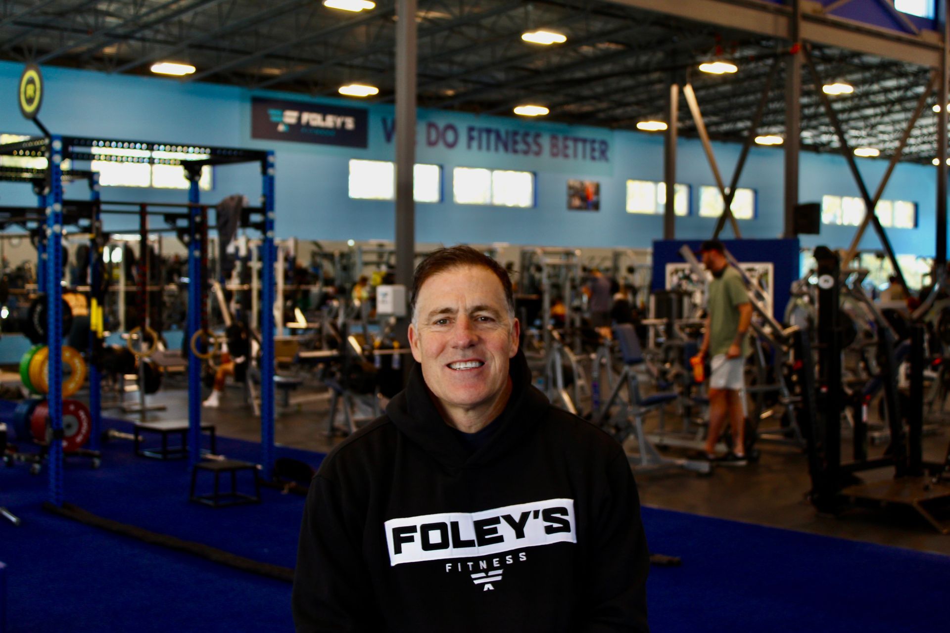 Man in black hoodie smiles at camera in a gym setting. The hoodie says 