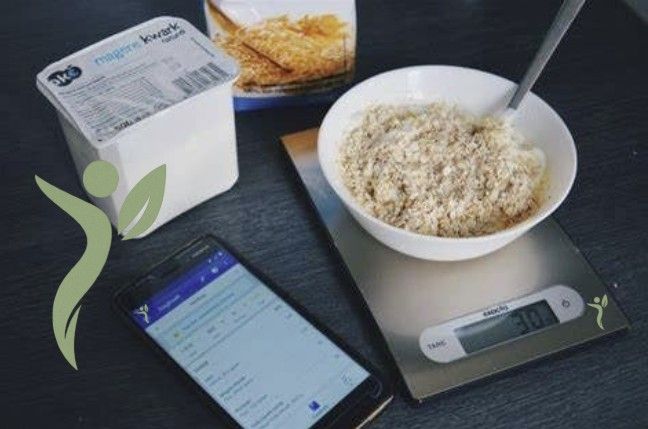 Een kom havermout op een weegschaal naast een bakje yoghurt, een voedselpakket en een telefoon met een app op een tafel.
