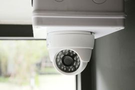 White security camera mounted indoors, facing downward near a window.