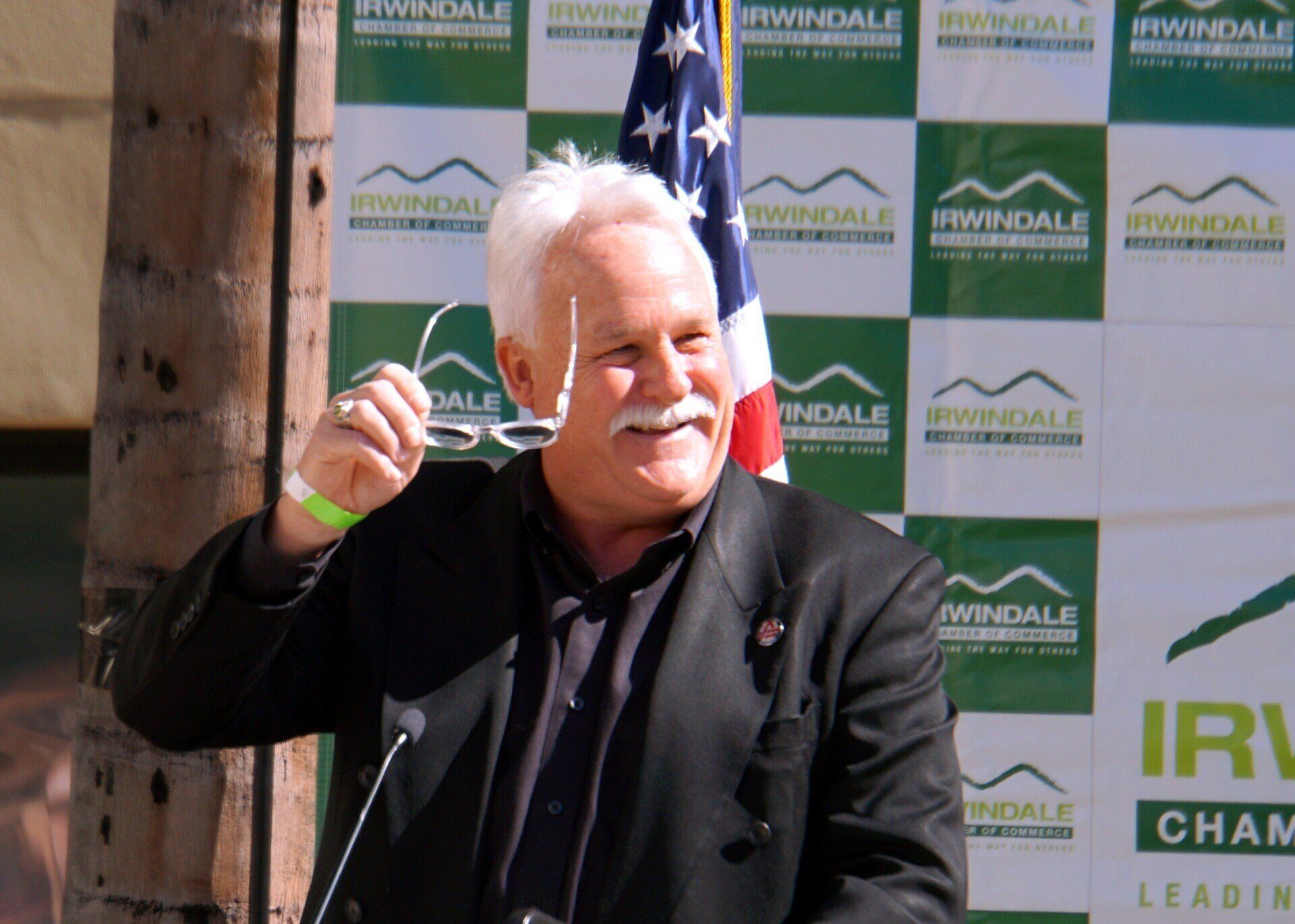 A man in a black suit stands in front of a podium with an american flag behind him