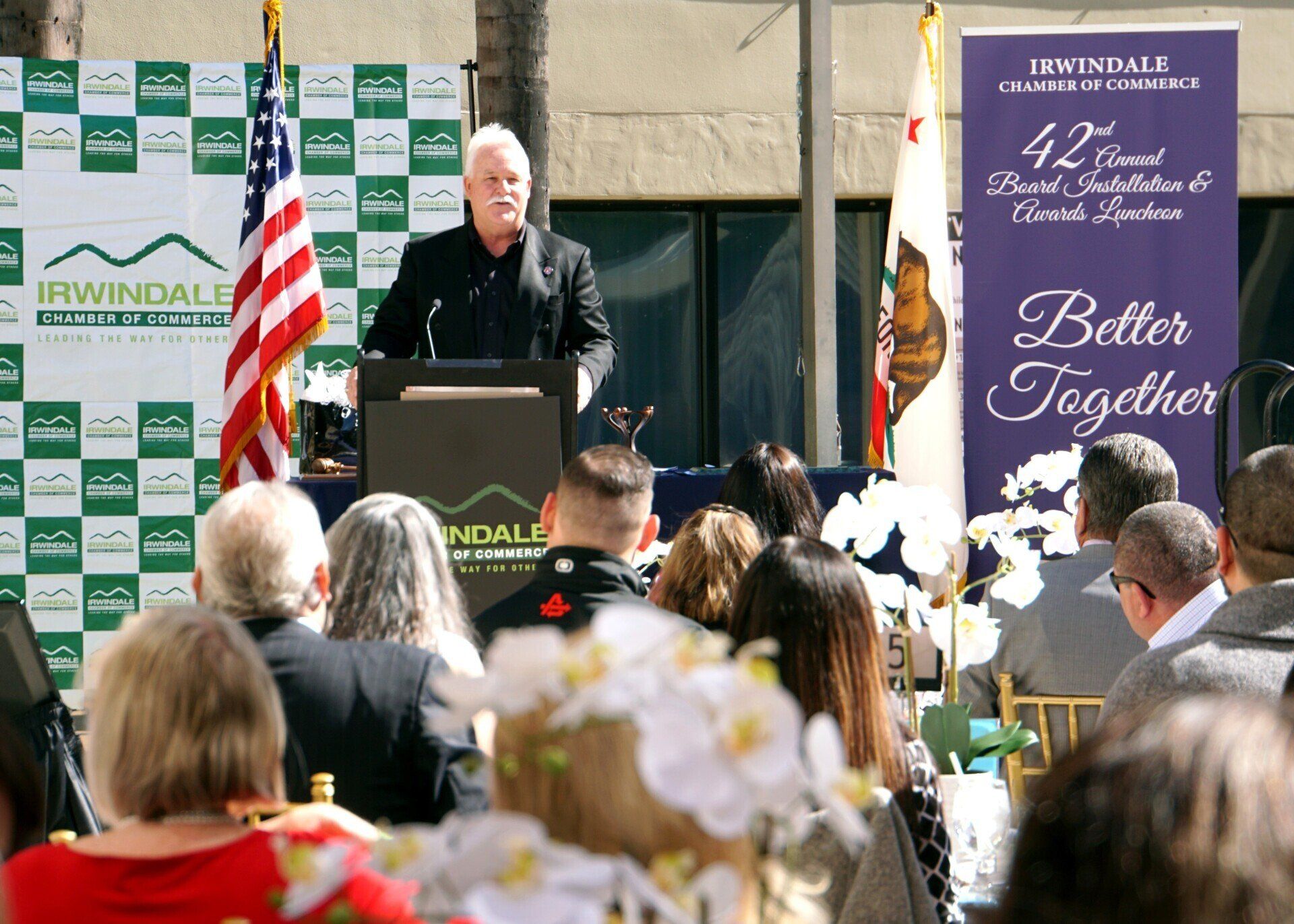A man is giving a speech in front of a crowd with a sign that says better together