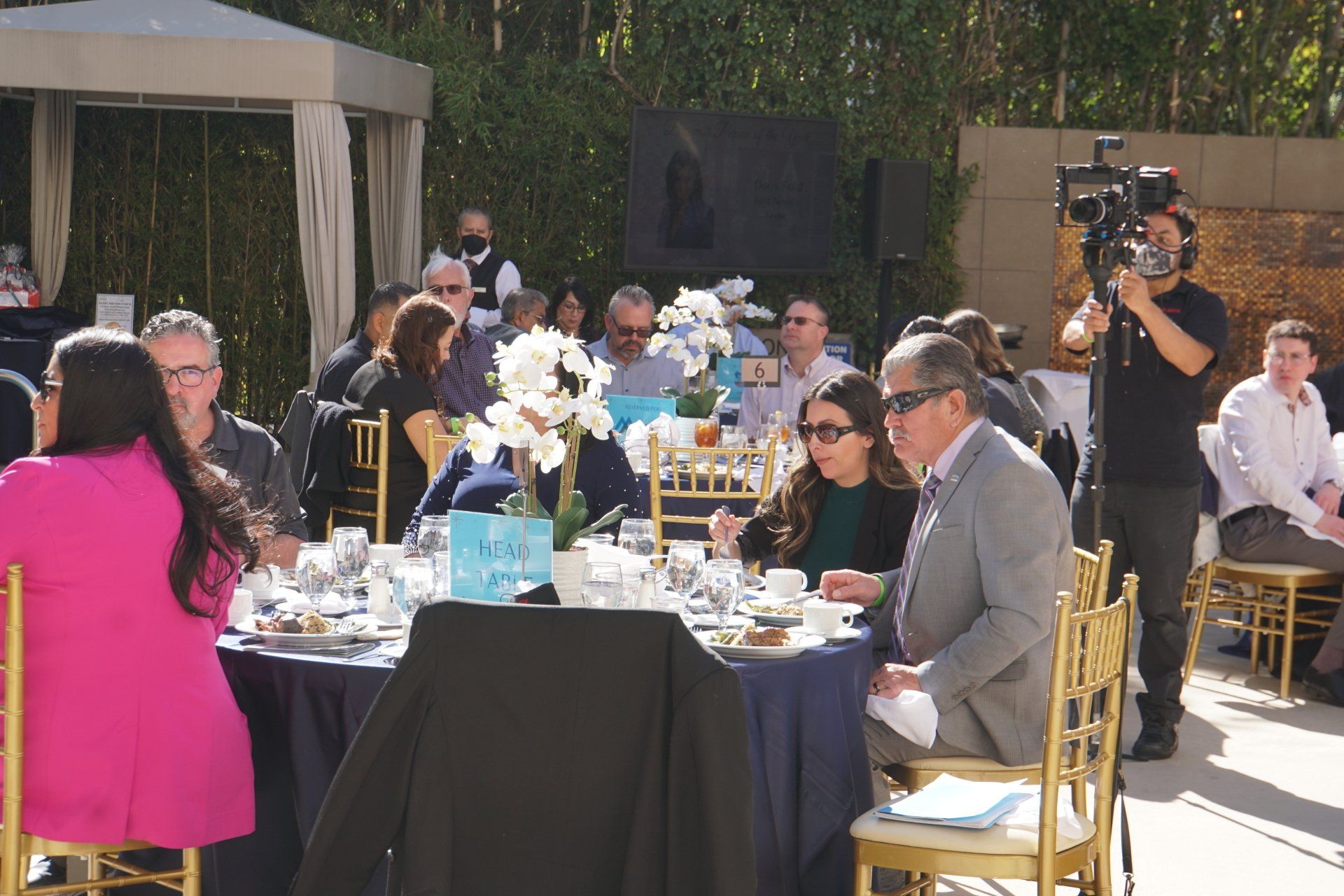 A group of people sitting at tables with a cameraman standing behind them