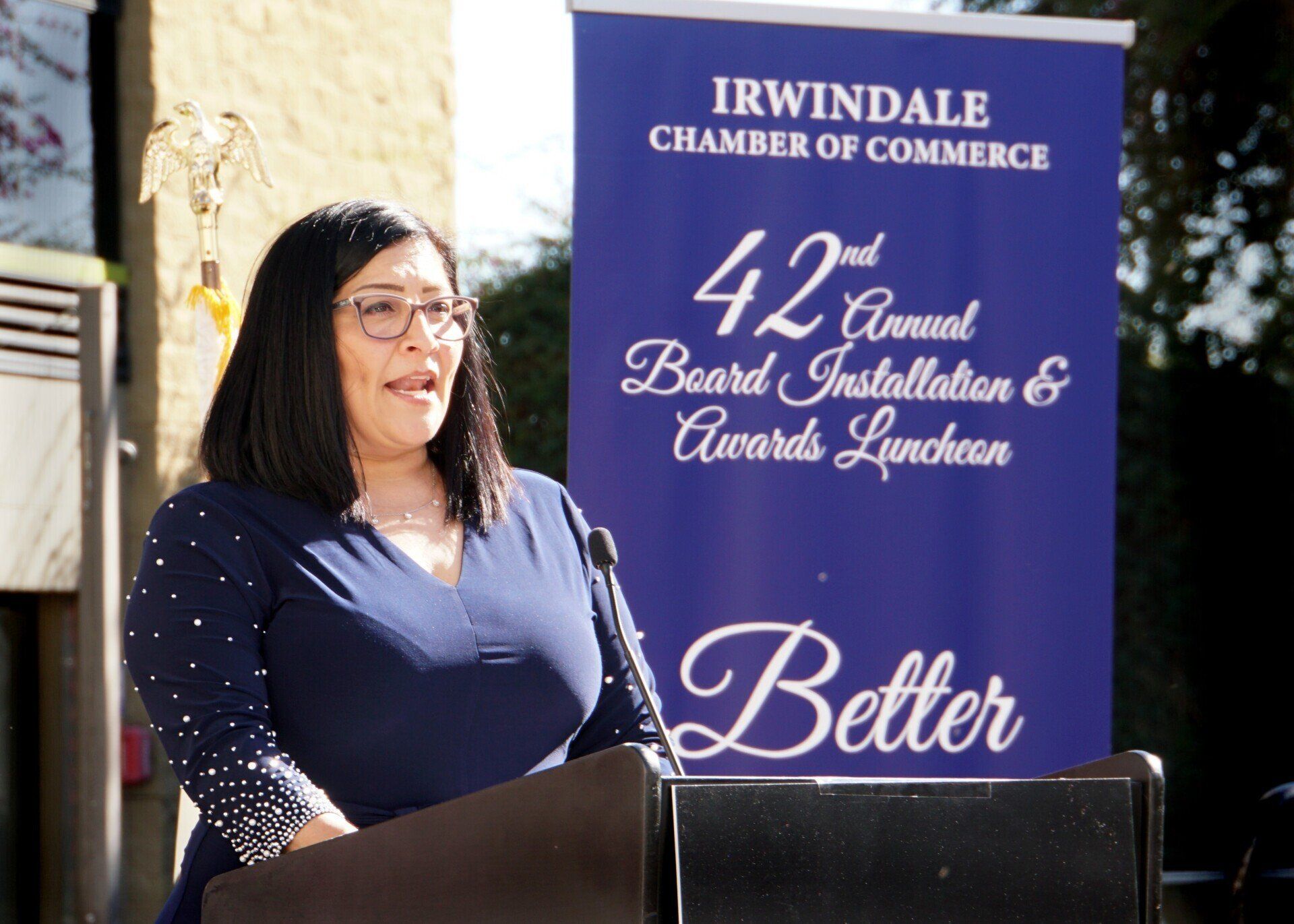 A woman stands at a podium in front of a banner for the 42nd annual board installation and awards luncheon