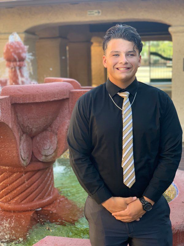 A man in a black shirt and tie is standing in front of a fountain.