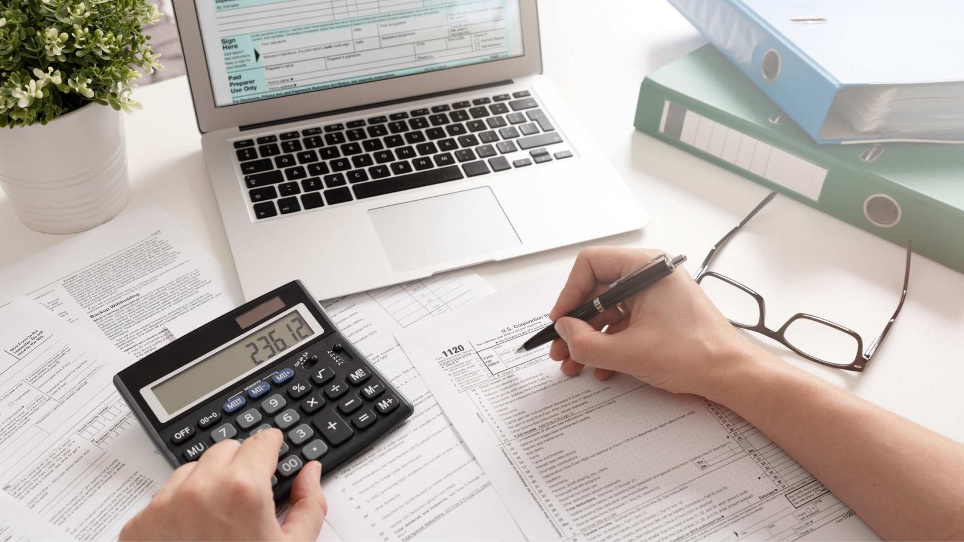 Person using a calculator and writing on papers at a desk with a laptop and glasses.