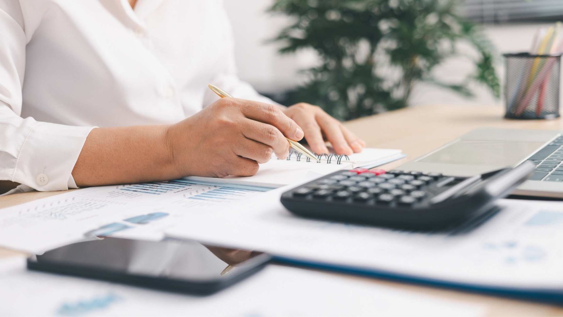 Person writing on paper beside a calculator and smartphone in a bright office workspace
