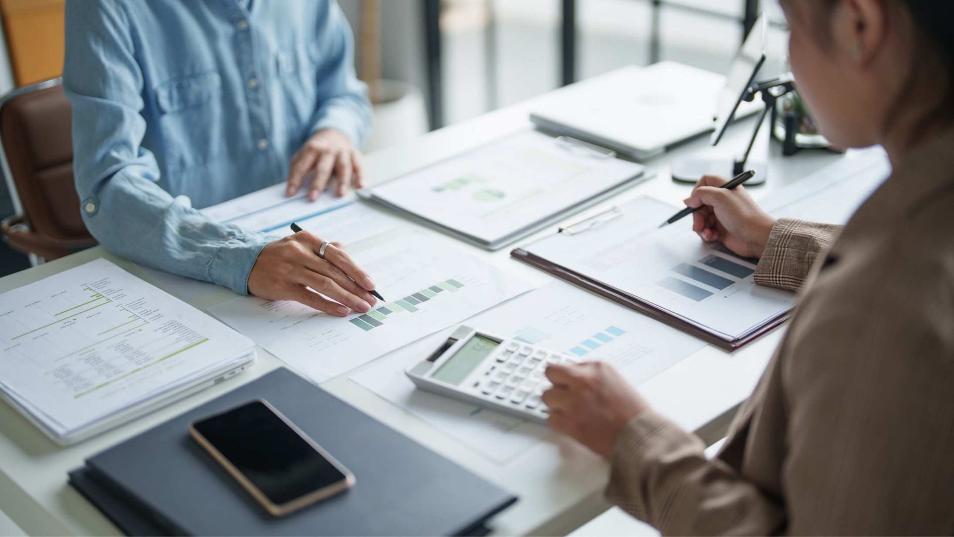 Two people reviewing financial charts and notes at a bright office table with a smartphone and calculator