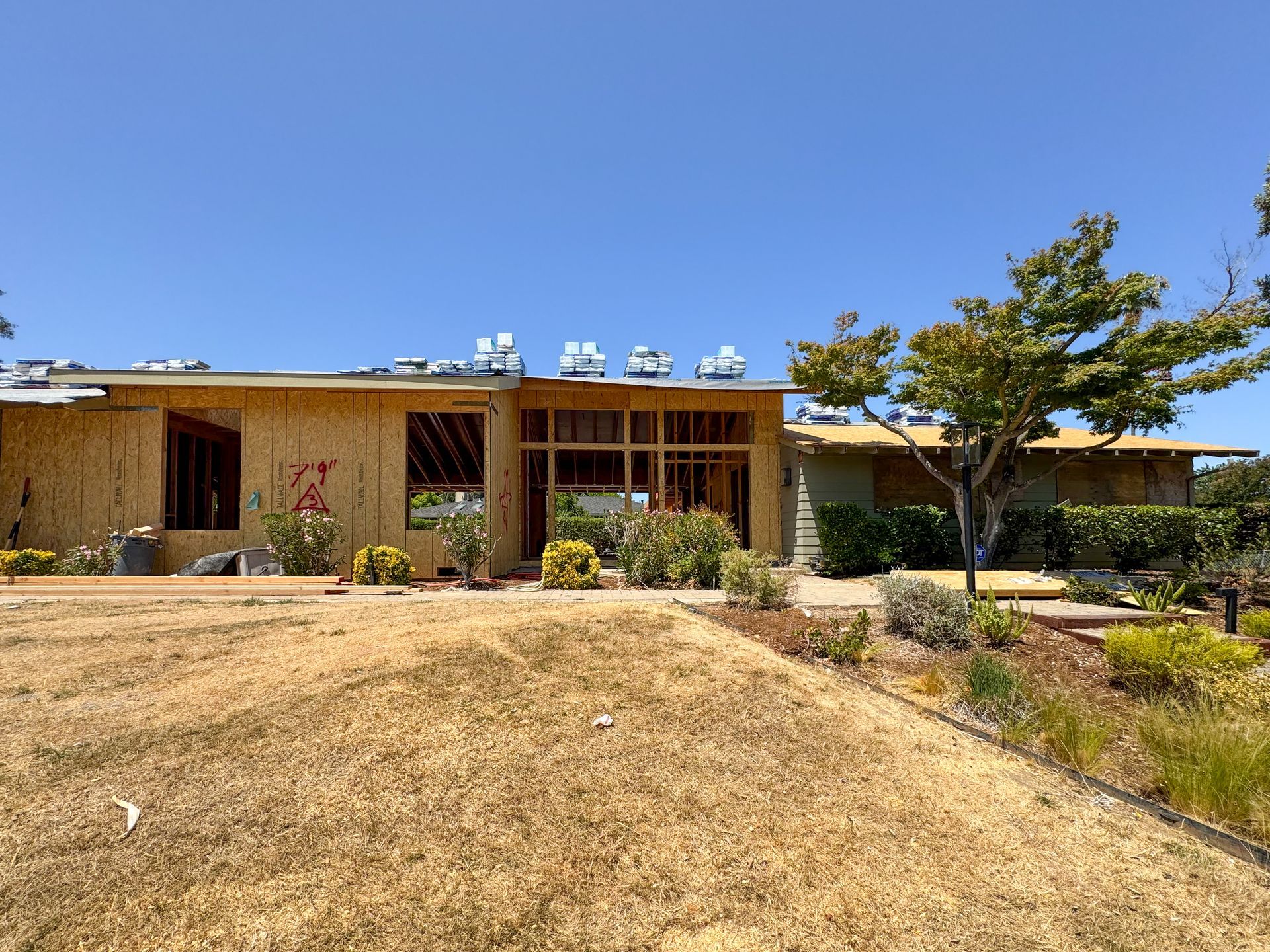 House under construction, with exposed wood framing, surrounded by dry grass and bushes on a sunny day.